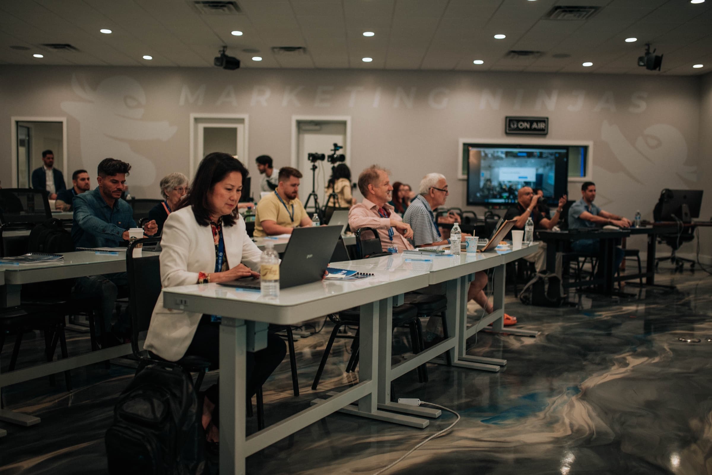 The AGM big room in session — attendees on laptops at glass desks, two production cameras rolling