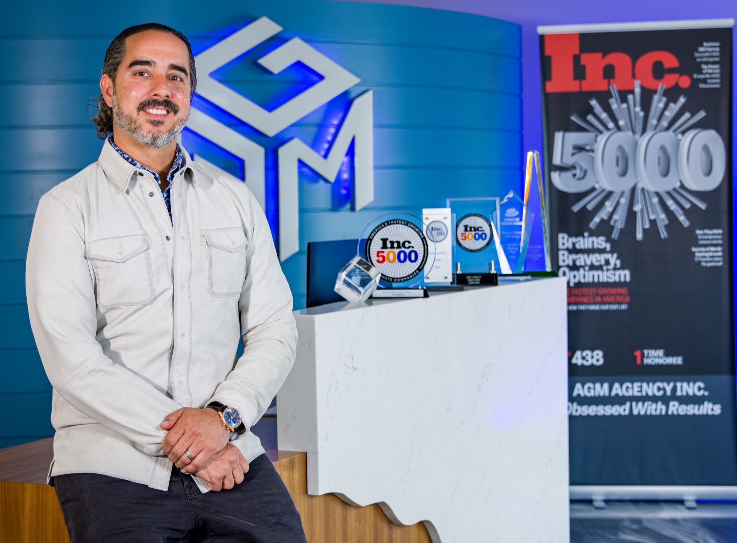Manuel Suarez beside the AGM logo wall with framed magazine covers and trophies