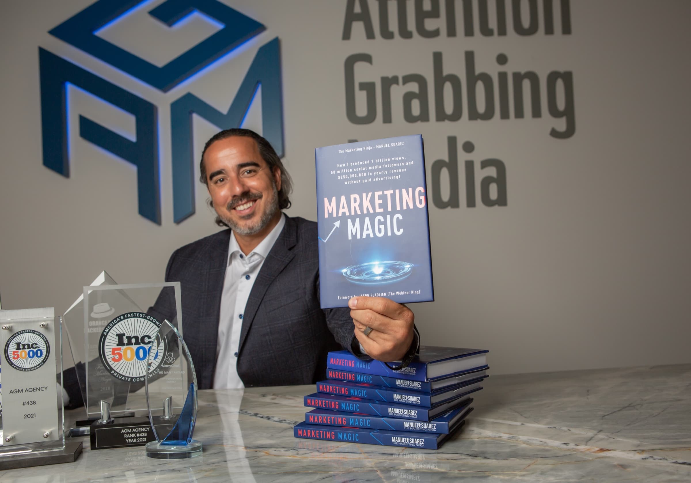 Manuel Suarez holding his book Marketing Magic in front of the Attention Grabbing Media wall, book stacks and trophies beside him