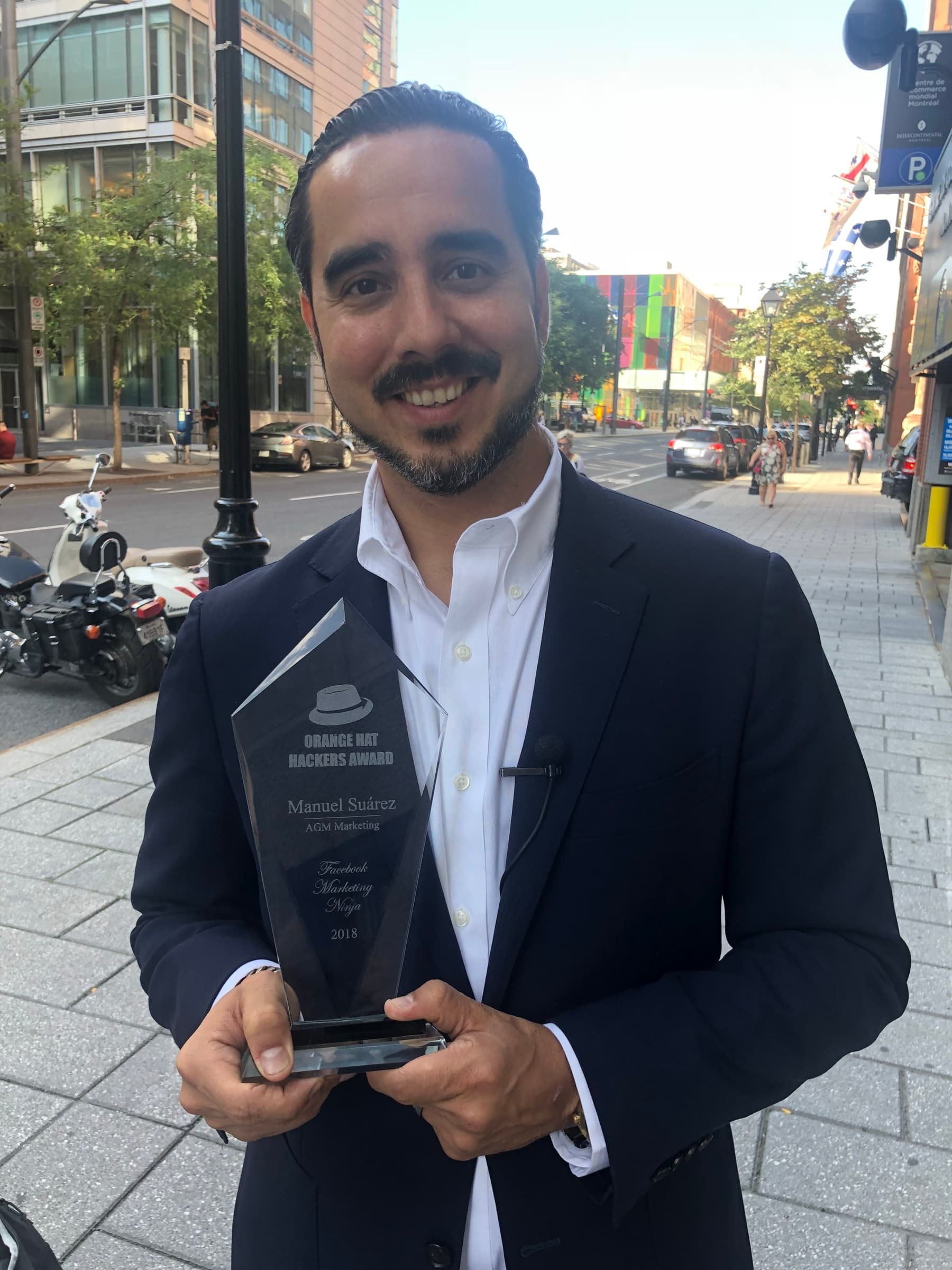Manuel Suarez in a suit holding the Orange Hacker trophy outdoors